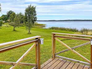 Een houten terras met een metalen reling, uitkijkend op een gazon met bomen en een waterpartij.