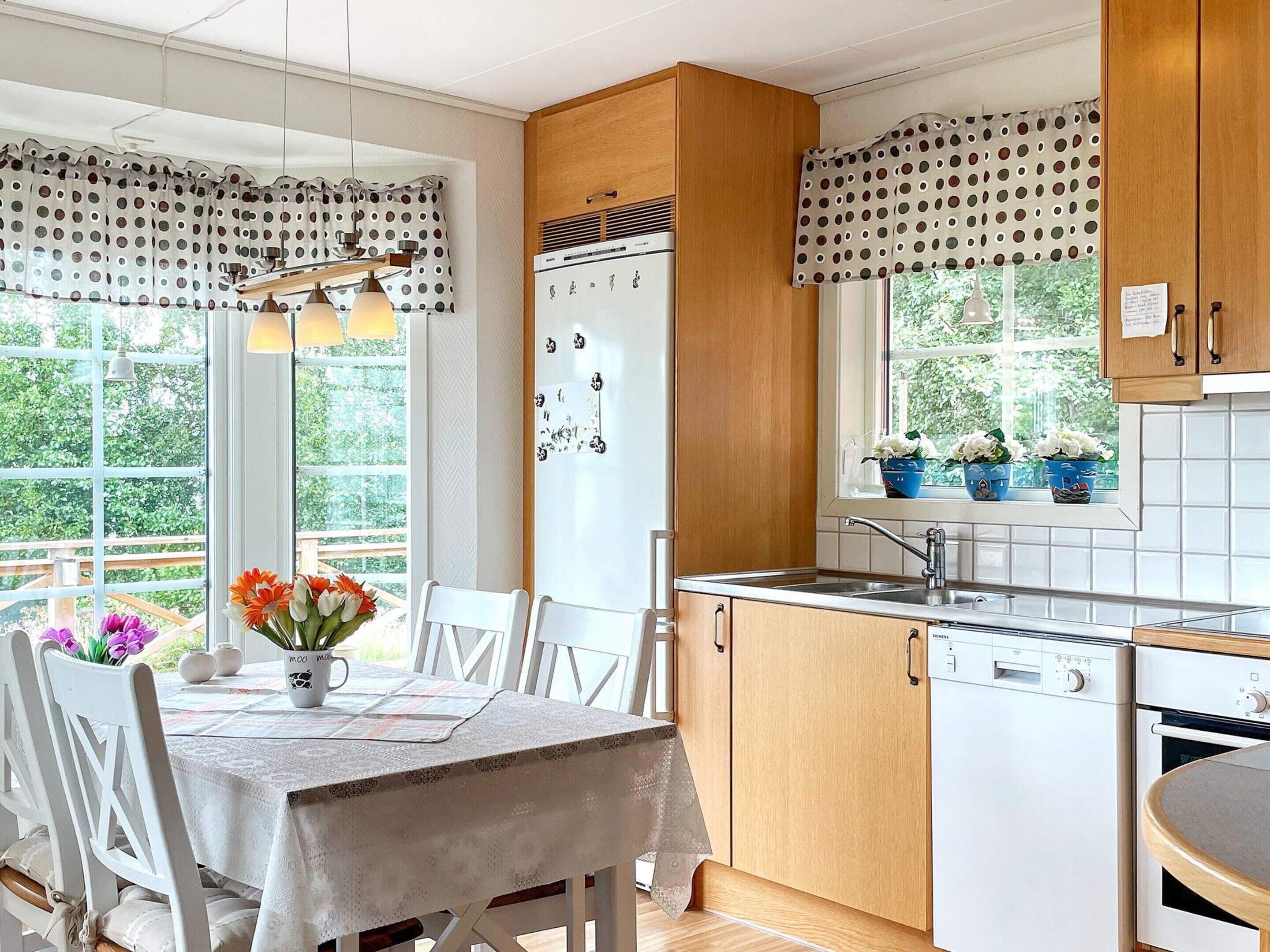 Una cocina con armarios de madera, un refrigerador blanco, un fregadero y una mesa de comedor con sillas. Hay plantas en macetas en el alféizar de la ventana y una cortina a lunares.