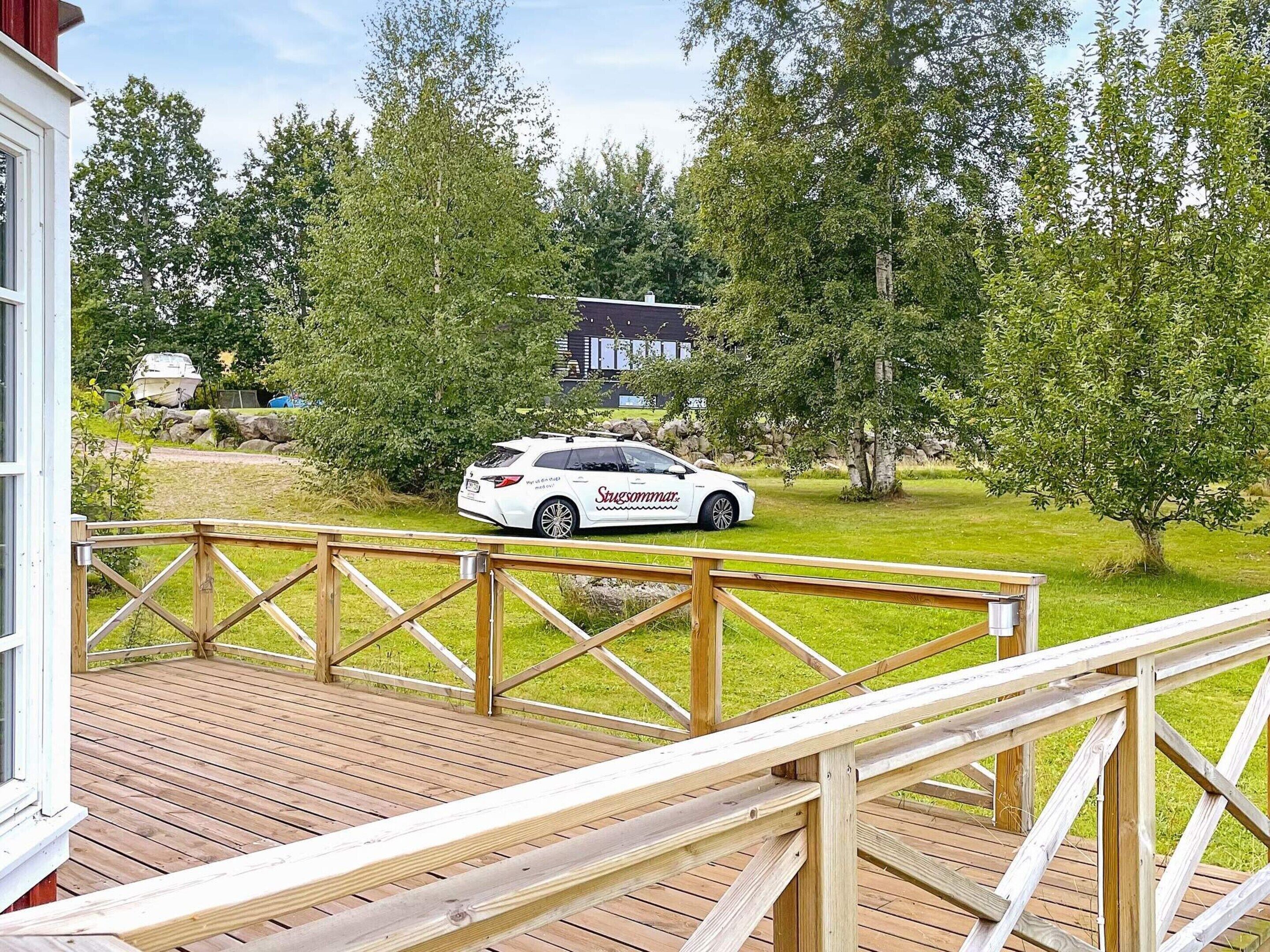 Un coche blanco aparcado en una terraza de madera, con una casa y árboles al fondo.