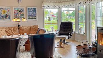 A cozy living room with a leather sofa, a dark armchair, a fireplace, and large windows offering a view of the outdoors.