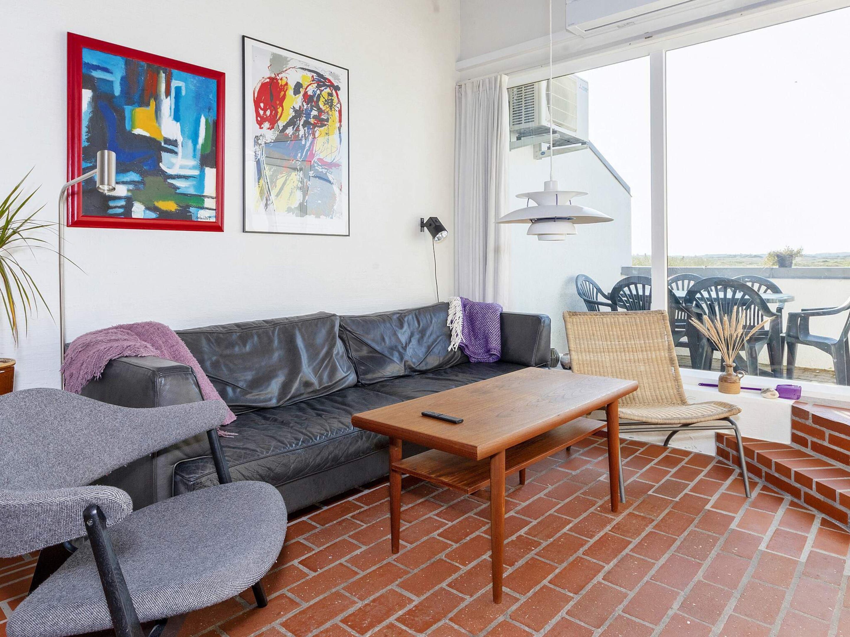 A living room with a leather sofa, a wooden coffee table, and two armchairs. There are two framed artworks on the wall, a ceiling fan, and a view of the outside through a large window.