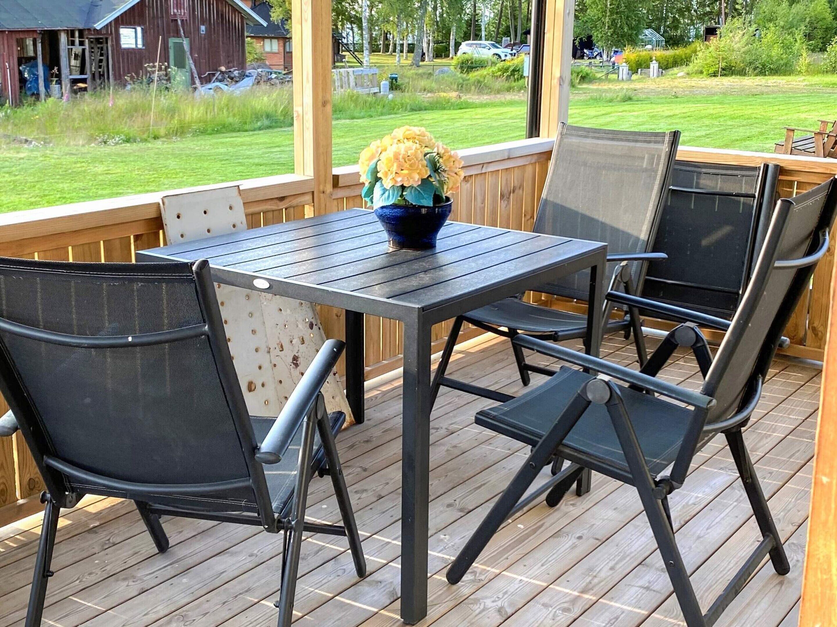 Un espace extérieur abrité, aménagé avec une table et des chaises, un vase de fleurs, et offrant une vue sur un champ verdoyant et des bâtiments.