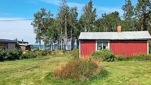 Une cabane en bois rouge, surmontée d’un toit en métal, entourée d’un champ verdoyant et de grands arbres.