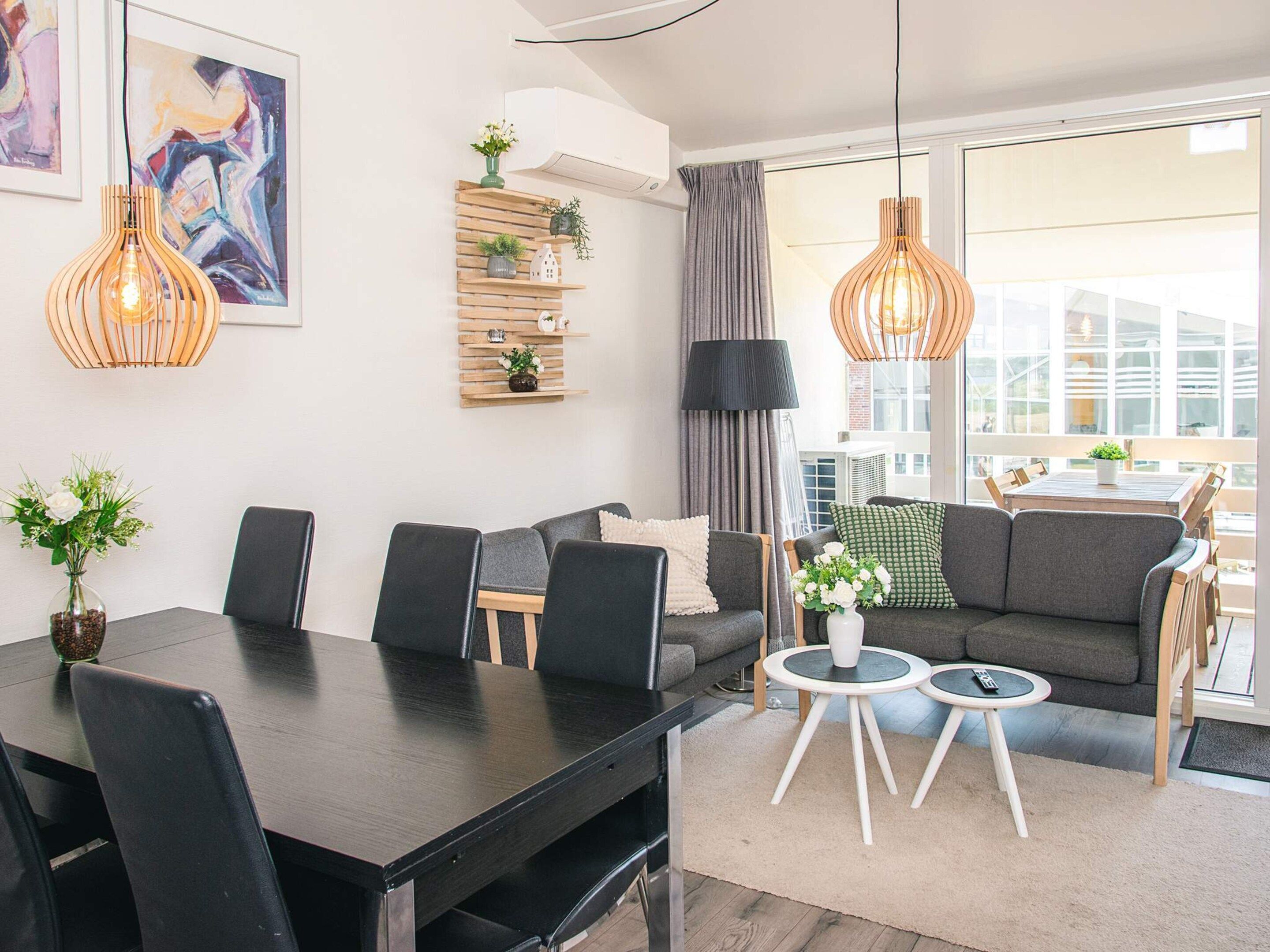 A modern living room with a dining table, sofas, and a coffee table. There are two pendant lights, a bookshelf, and a large window with curtains.