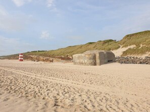 A sandy beach with a large concrete structure, a red and white striped marker, and a grassy dune in the background. - 6 Person Holiday Home in Ringkobing (Ringkobing)