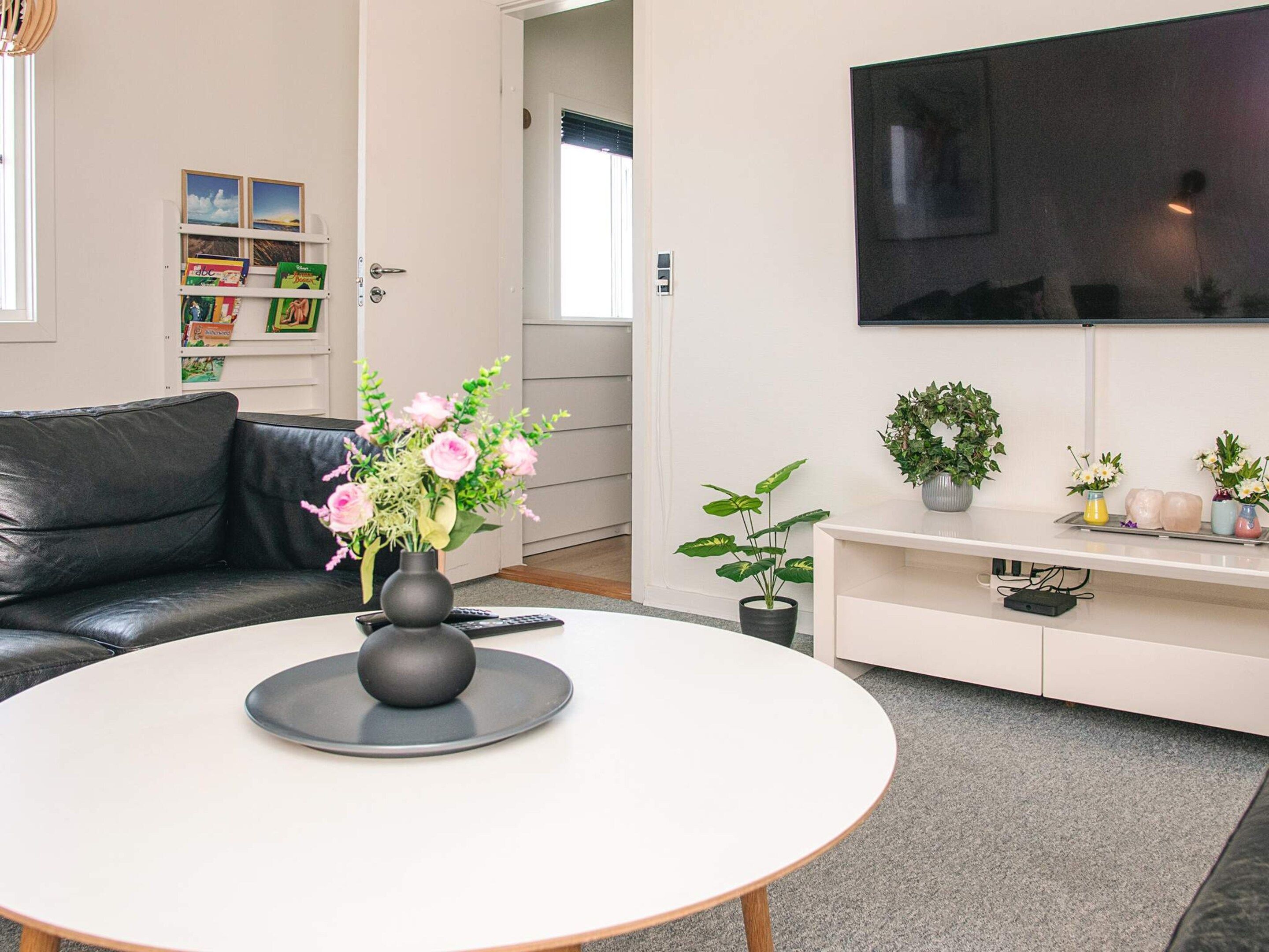 Ein modernes Wohnzimmer mit einem schwarzen Ledersofa, einem runden weißen Couchtisch mit Blumenvase, einem flachbildfernseher an der Wand und einem Bücherregal mit Kinderbüchern.