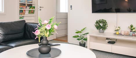 Uma sala de estar moderna com um sofá de couro preto, uma mesa de centro redonda branca com um vaso de flores, uma TV de tela plana na parede e uma estante com livros infantis.
