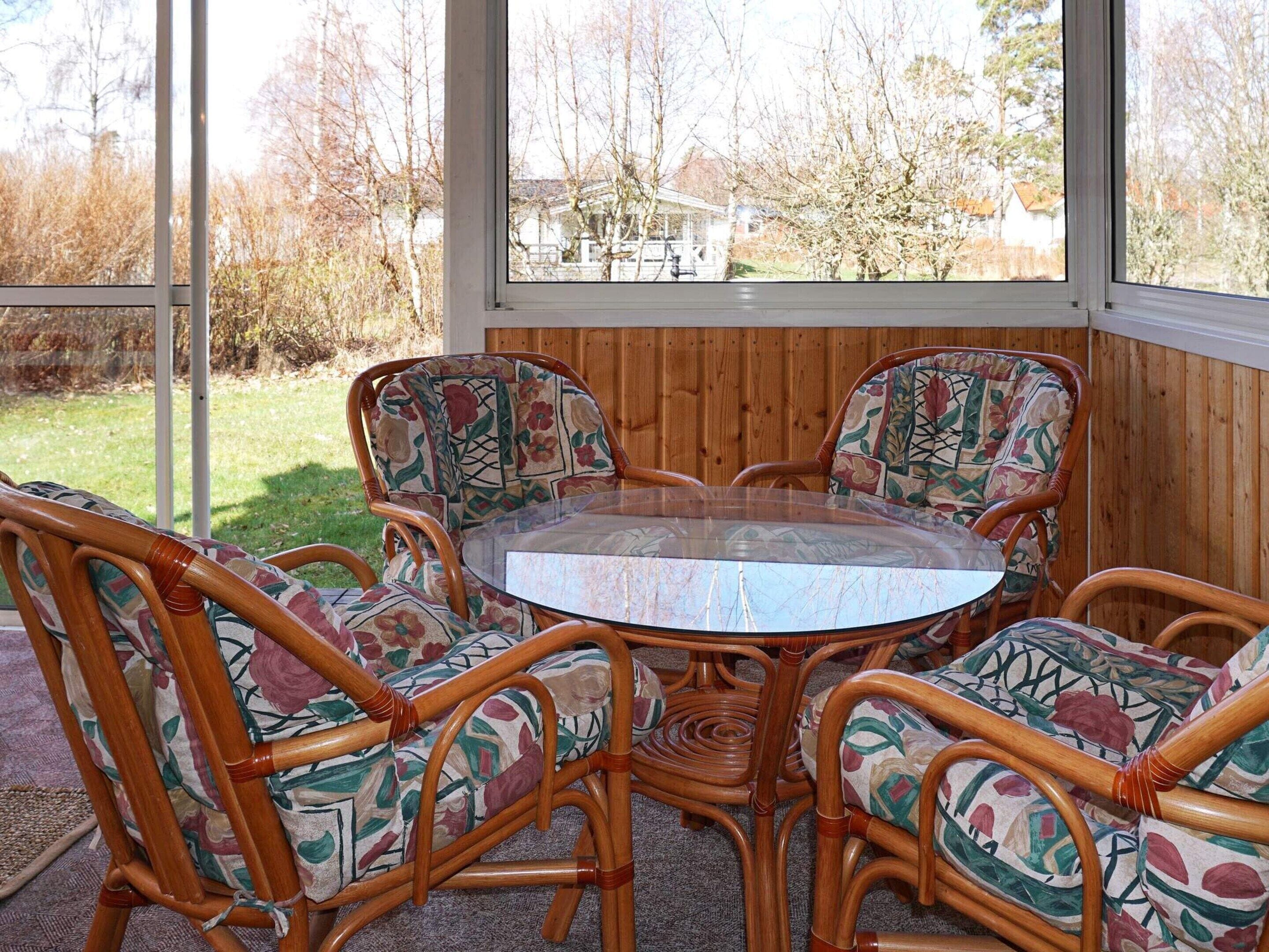 A round glass table surrounded by four wicker chairs with floral upholstery, set against a wooden paneled wall with a view of a residential area.