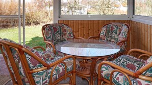 Une table ronde en verre, entourée de quatre chaises en osier à motifs floraux, est placée devant un mur lambrissé, offrant une vue sur un quartier résidentiel.