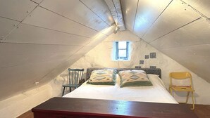 Une petite chambre mansardée, chaleureuse et accueillante, avec un plafond en pente, une commode en bois ornée de motifs sculptés, un lit simple avec des coussins à motifs, une petite fenêtre et une chaise.