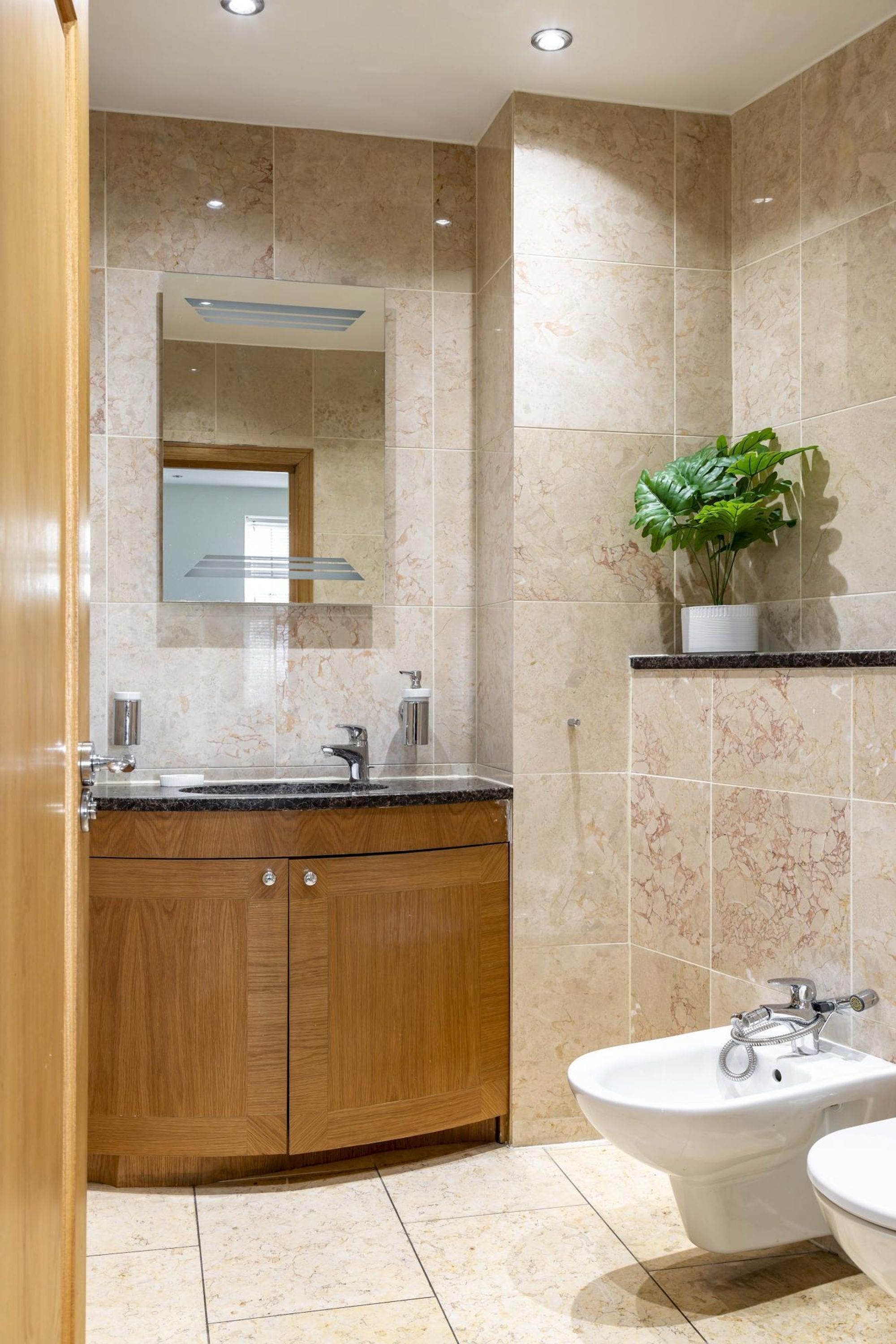 A modern bathroom with a marble-tiled wall, a wooden vanity, a bidet, and a potted plant.