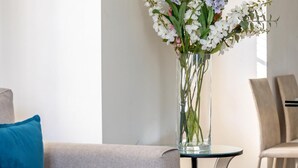 A modern living room with a grey sofa, a glass coffee table, and a vase of flowers. There are framed pictures on the wall and a chair in the background.