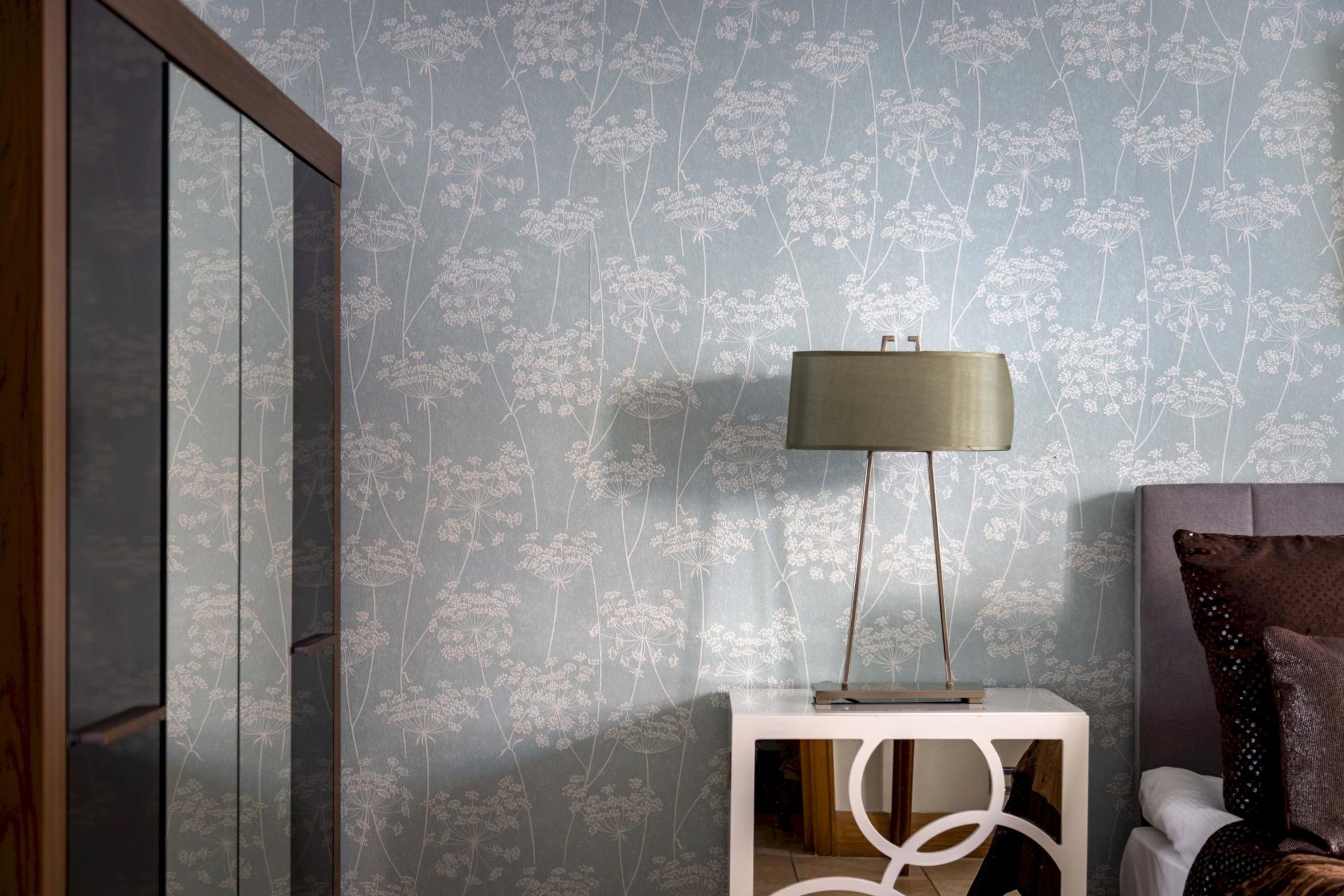 Una habitación con papel tapiz estampado, una mesita de noche con lámpara y una cama con cabecero.