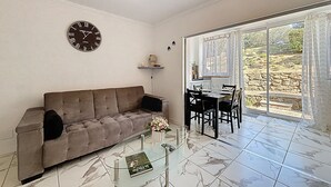 Un salon comprenant un canapé, une table basse en verre, une table à manger avec des chaises, une horloge et une vue sur un jardin, visible à travers une porte coulissante.