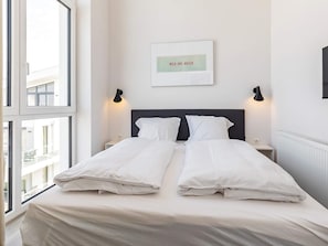 Una habitación de hotel con una cama, dos mesitas de noche, un ventanal y un cuadro colgado en la pared.