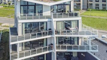 Un edificio moderno de varios pisos con balcones de vidrio, mobiliario exterior y un techo plano con vegetación.