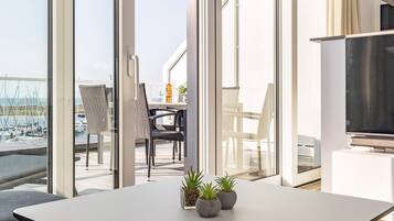 Un interior moderno con balcón que da a un puerto deportivo, con zona de comedor con sillas y mesa, y un televisor de pantalla plana sobre un soporte.