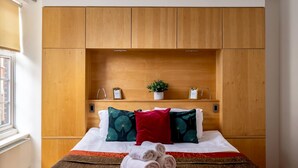 Hotelkamer met houten hoofdeinde, een bed met witte lakens, twee decoratieve kussens, een rood accentkussen, een kleine potplant en een set handdoeken op het bed.