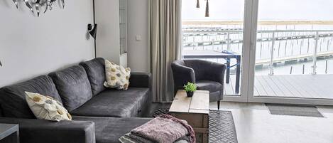 A modern living room with a grey sofa, a small table, a plant, and a view of a pier through a glass door.