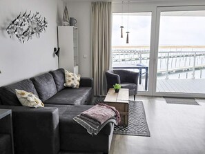 A modern living room with a grey sofa, a small table, a plant, and a view of a pier through a glass door. - 5 Star Holiday Home in Wendtorf (Wendtorf)