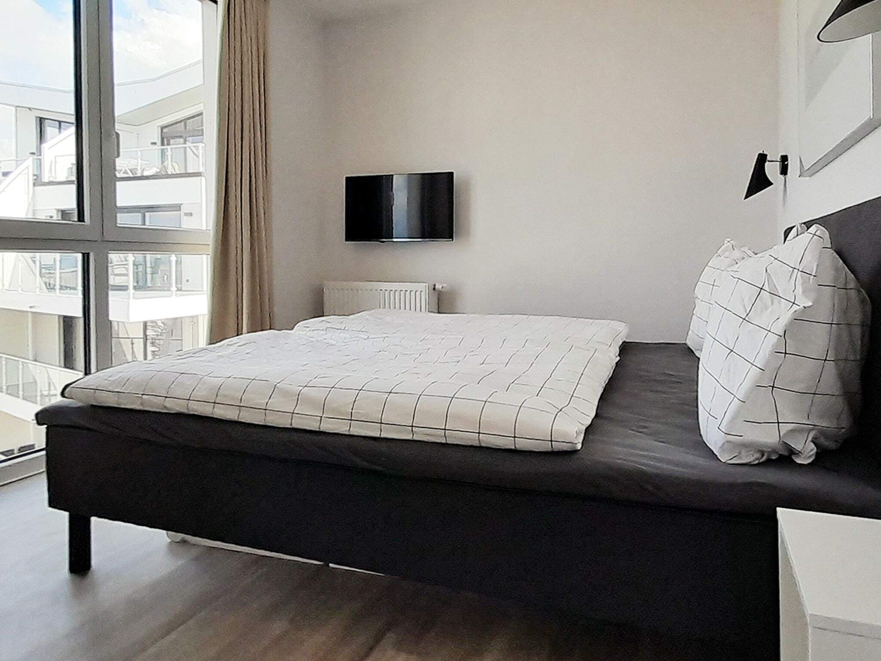 Une chambre moderne avec un lit, un téléviseur à écran plat et une vue sur un immeuble, visible à travers de grandes fenêtres.
