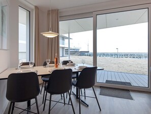 Un coin repas avec une table dressée pour quatre personnes, de grandes fenêtres donnant sur la plage et un luminaire suspendu moderne.