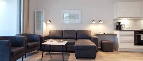 A modern living room with a sofa, armchairs, and a coffee table. There is a kitchen area with a coffee machine and cabinets.