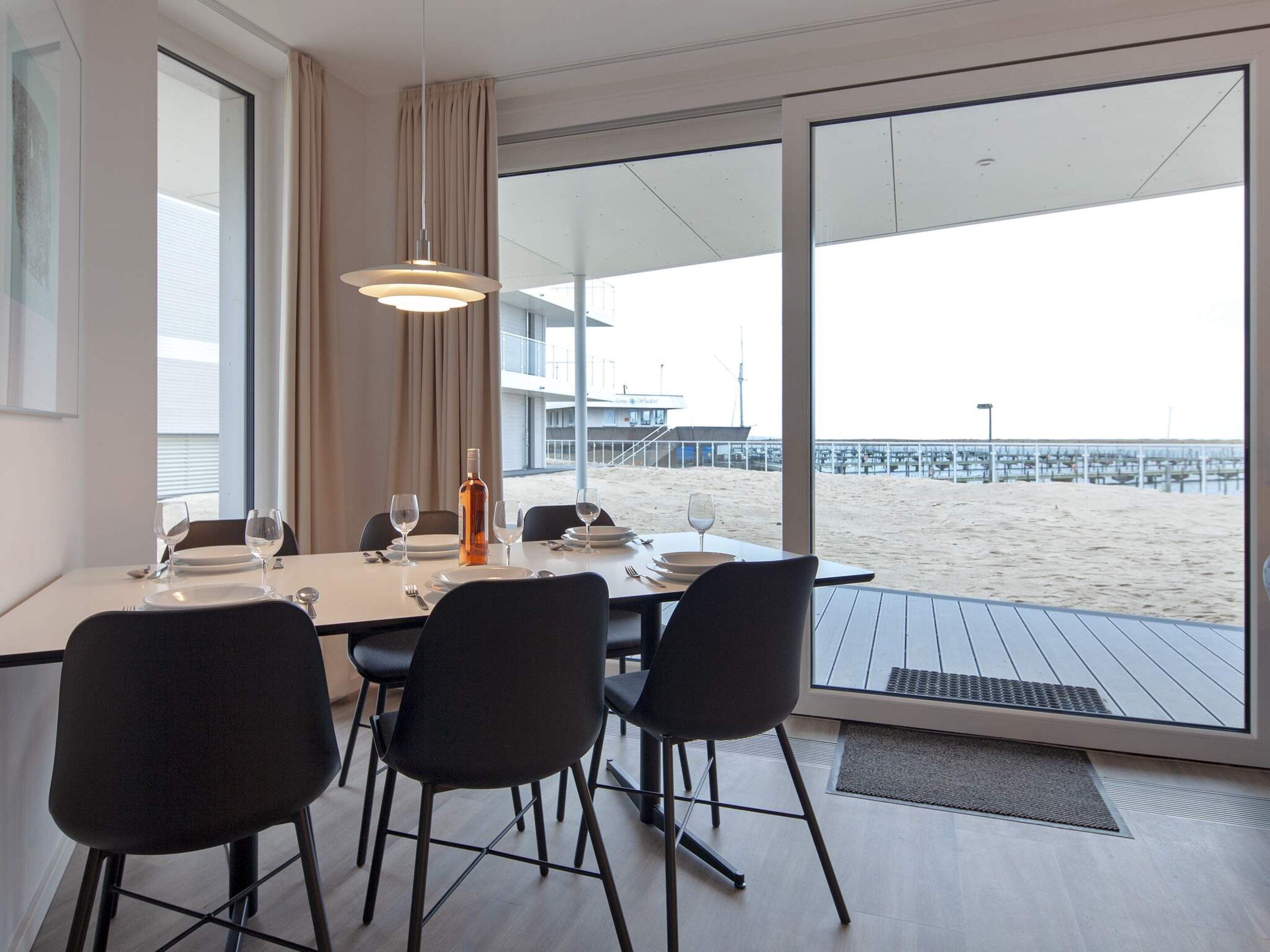A dining area with a table set for four, a bottle of wine, and a view of the beach through large windows.