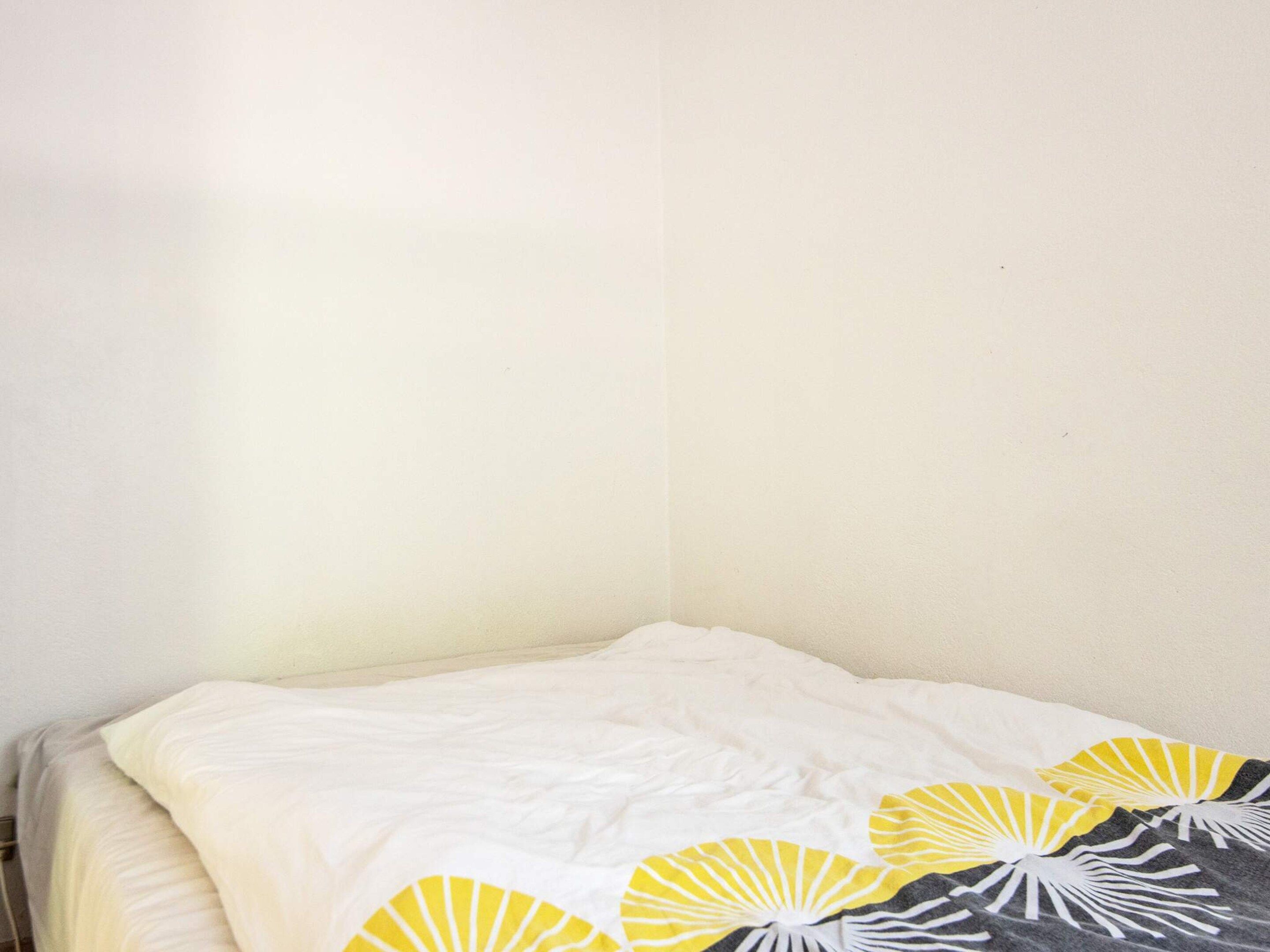 A bed with a white sheet and a blanket, a plain wall, and a corner of a room.