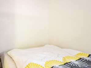 Un lit avec un drap blanc et une couverture, un mur uni et un coin de pièce.