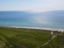 Vista aérea de uma área costeira com praia, campos gramados e céu claro.