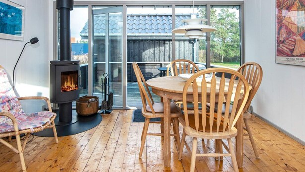 A cozy room with a wood-burning stove, a dining table with chairs, and a large window overlooking a garden. - 6 Person Holiday Home in Glesborg (Glesborg)