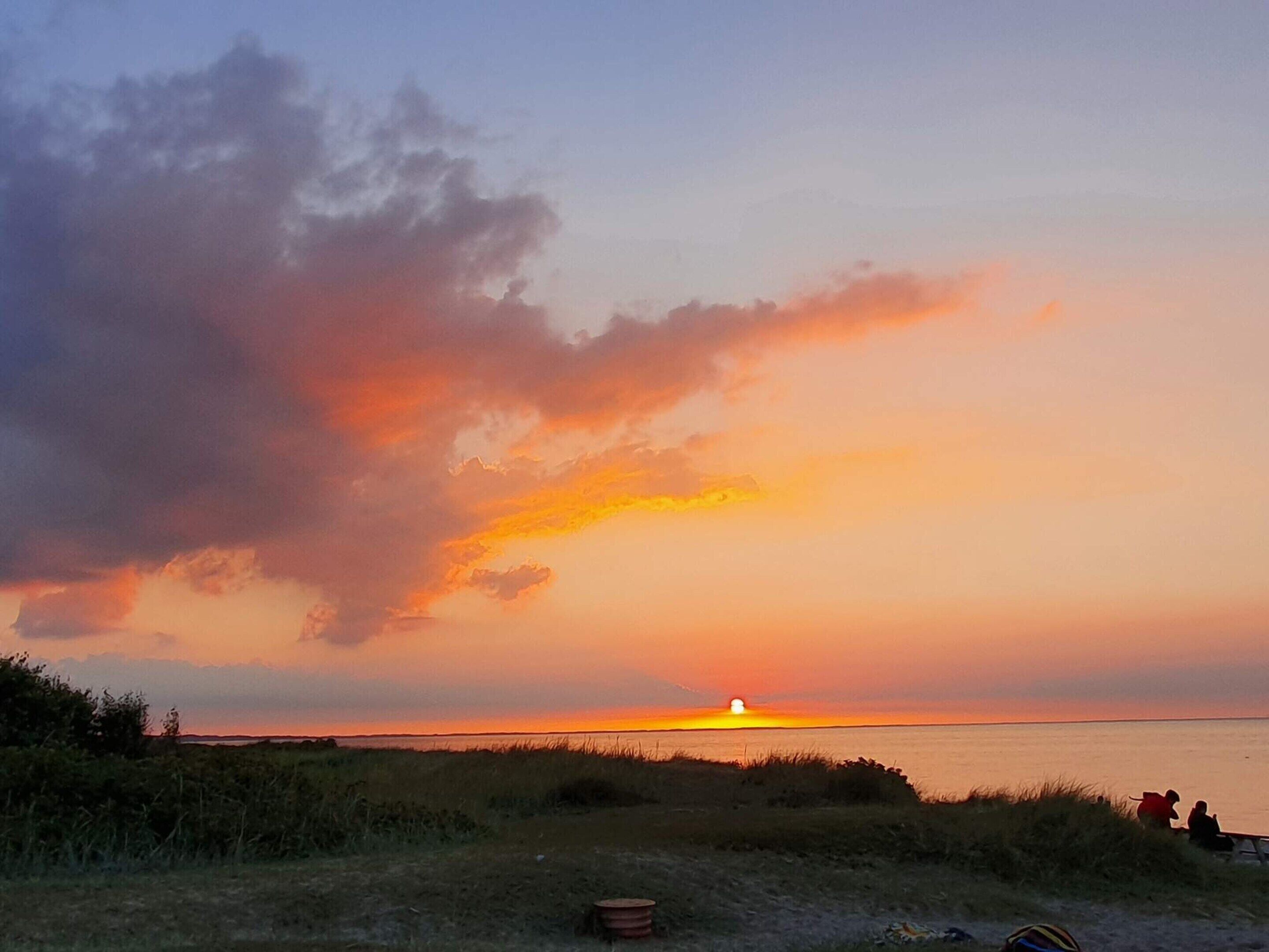 A sunset over a body of water with a grassy foreground and a few people sitting on the shore.