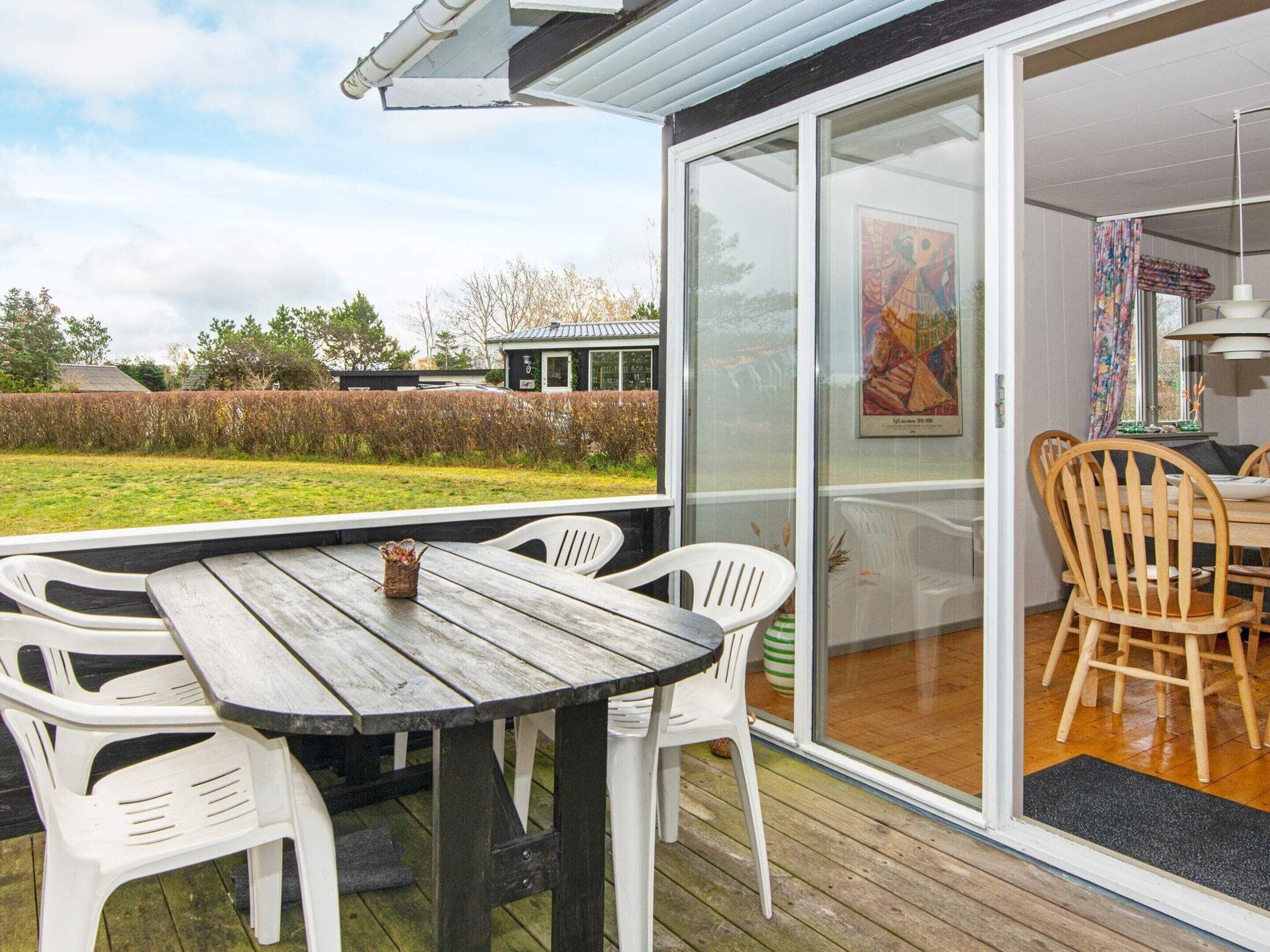 A wooden deck with a table and chairs, a glass door leading to an indoor area, and a view of a garden and a house in the background.