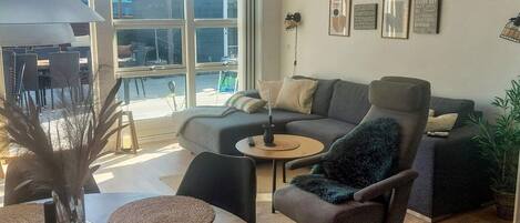 A modern living room with a sofa, armchair, and coffee table. There are framed pictures on the wall and a large window providing natural light.