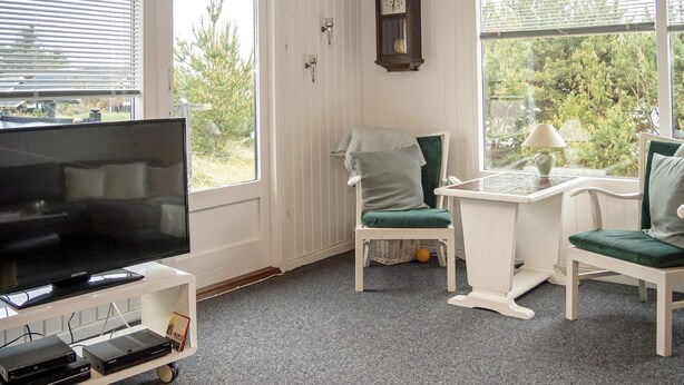 A room with a television on a stand, a clock on the wall, and two chairs with cushions. - 6 Person Holiday Home in Thisted (Thisted)