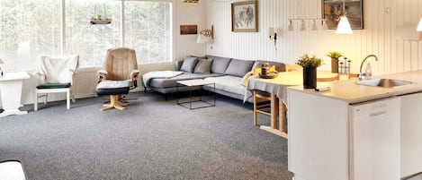 A modern living room with a grey sofa, a white dining area, and a kitchen with a sink.