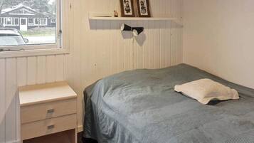 A room with a bed, a small bedside table, a shelf with framed pictures, and a window with a view of a house and a car.