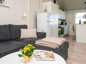 Uma sala de estar moderna com um sofá cinzento, uma mesa de centro branca com uma revista e flores, e uma cozinha ao fundo.