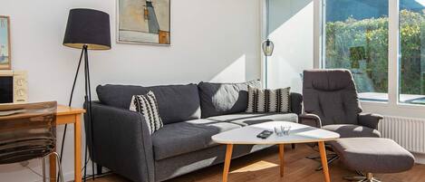 Un salon moderne avec un canapé gris, une petite table basse, un fauteuil gris et une grande fenêtre donnant sur un paysage verdoyant et montagneux.