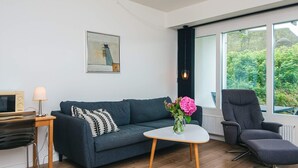 A modern living room with a sofa, a coffee table, a chair, a lamp, a TV, and a window with a view of greenery. - 4 Star Holiday Home in Ringkobing (Ringkobing)