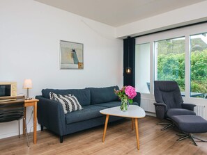 Ein modernes Wohnzimmer mit einer Couch, einem Couchtisch, einem Sessel, einer Lampe, einem Fernseher und einem Fenster mit Blick auf Grünflächen.