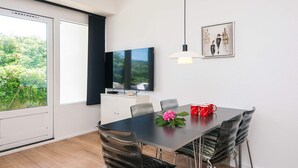 A modern dining room with a large table, chairs, a TV, and a view of greenery. - 4 Star Holiday Home in Ringkobing (Ringkobing)