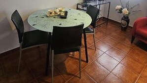 Et spiseområde med et rundt bord, stole, en laptop på et stativ, en vase med blomster og et vindue med gardiner.
