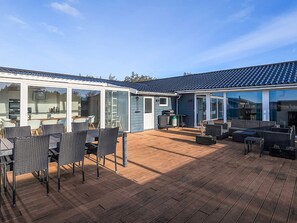 A modern patio area with a wooden deck, outdoor furniture, and a glass-enclosed kitchen. - 5 Star Holiday Home in Lokken (Lokken)