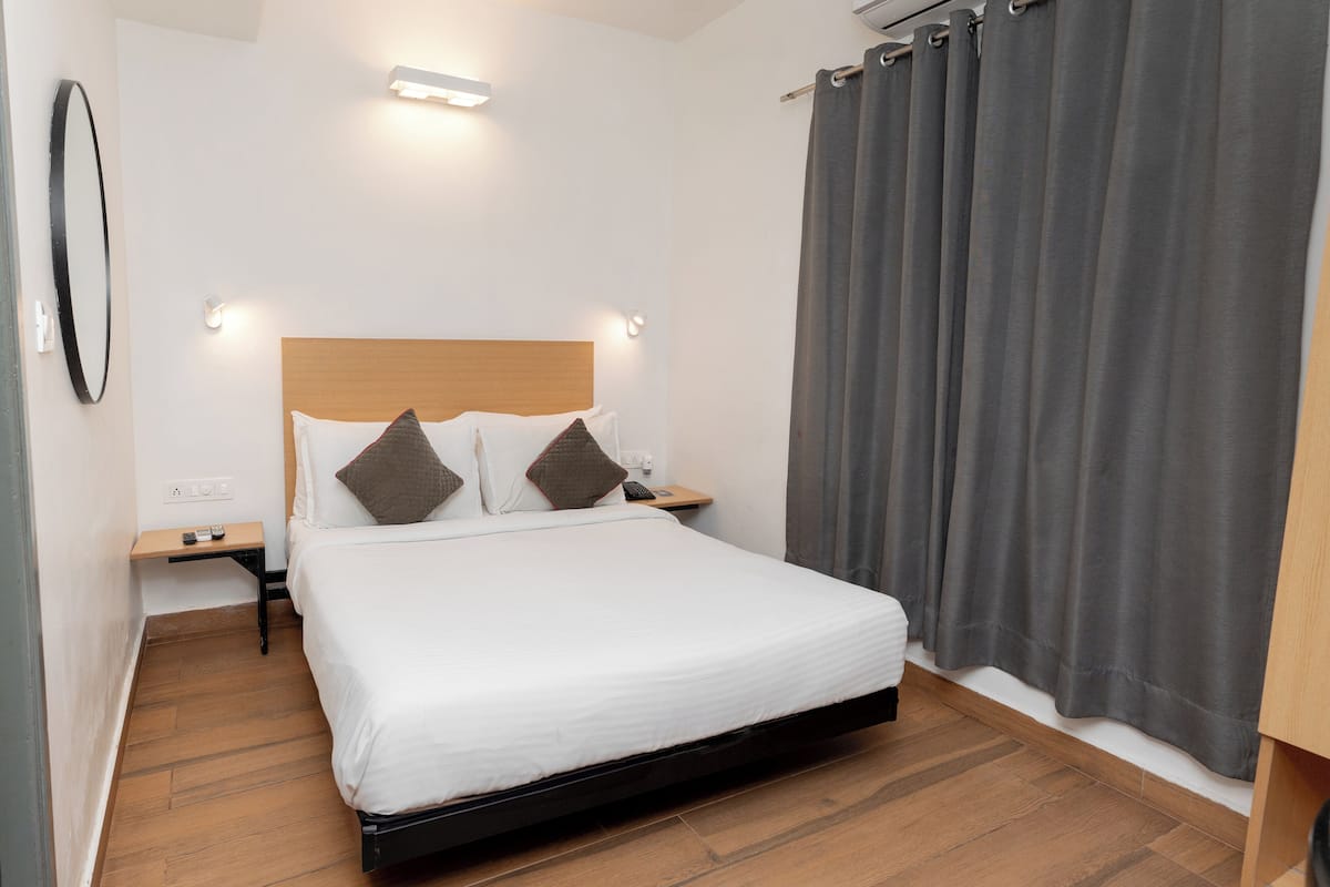 Una cama bien hecha con sábanas blancas y dos almohadas, una mesita de noche con control remoto y una cortina gris en la ventana.