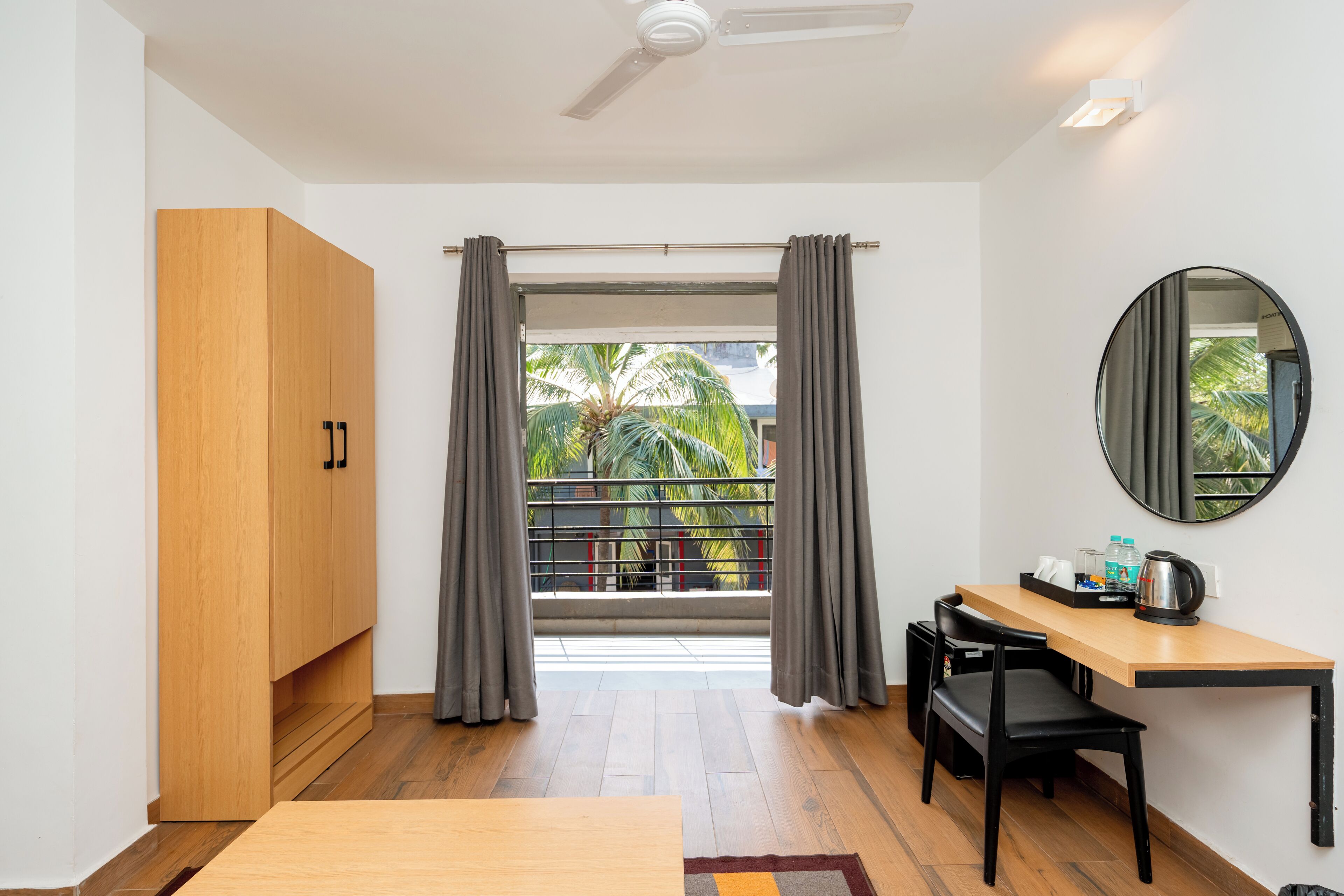 A room with a wooden wardrobe, a desk with a chair, a round mirror, and a view of a balcony with curtains.
