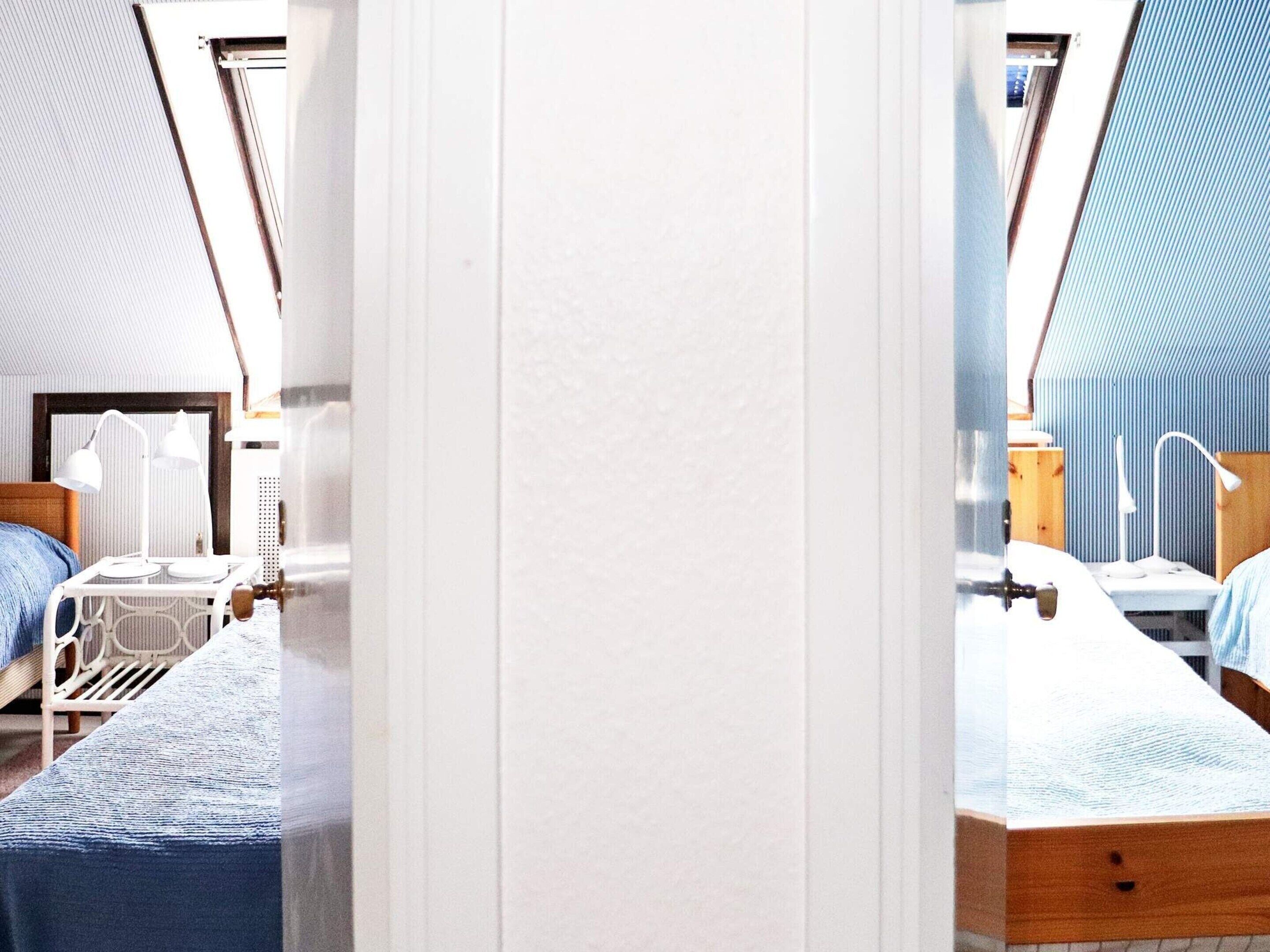 A room with a sloped ceiling, a bed with a wooden headboard, a bedside table with a lamp, and a window with a view of the outside.