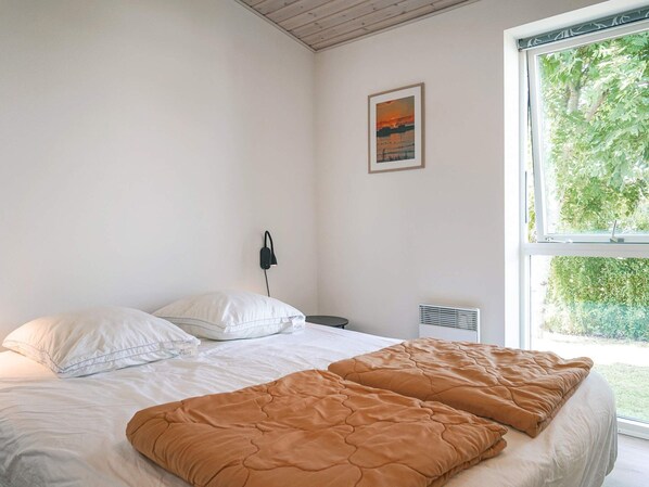 A bedroom with a bed, white pillows, a brown blanket, a nightstand, a wall-mounted lamp, and a window with a view of greenery.