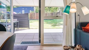 Uma sala de estar moderna com uma porta de vidro deslizante que dá para um pátio. Há um sofá com almofada, uma mesa com cadeiras e um abajur.